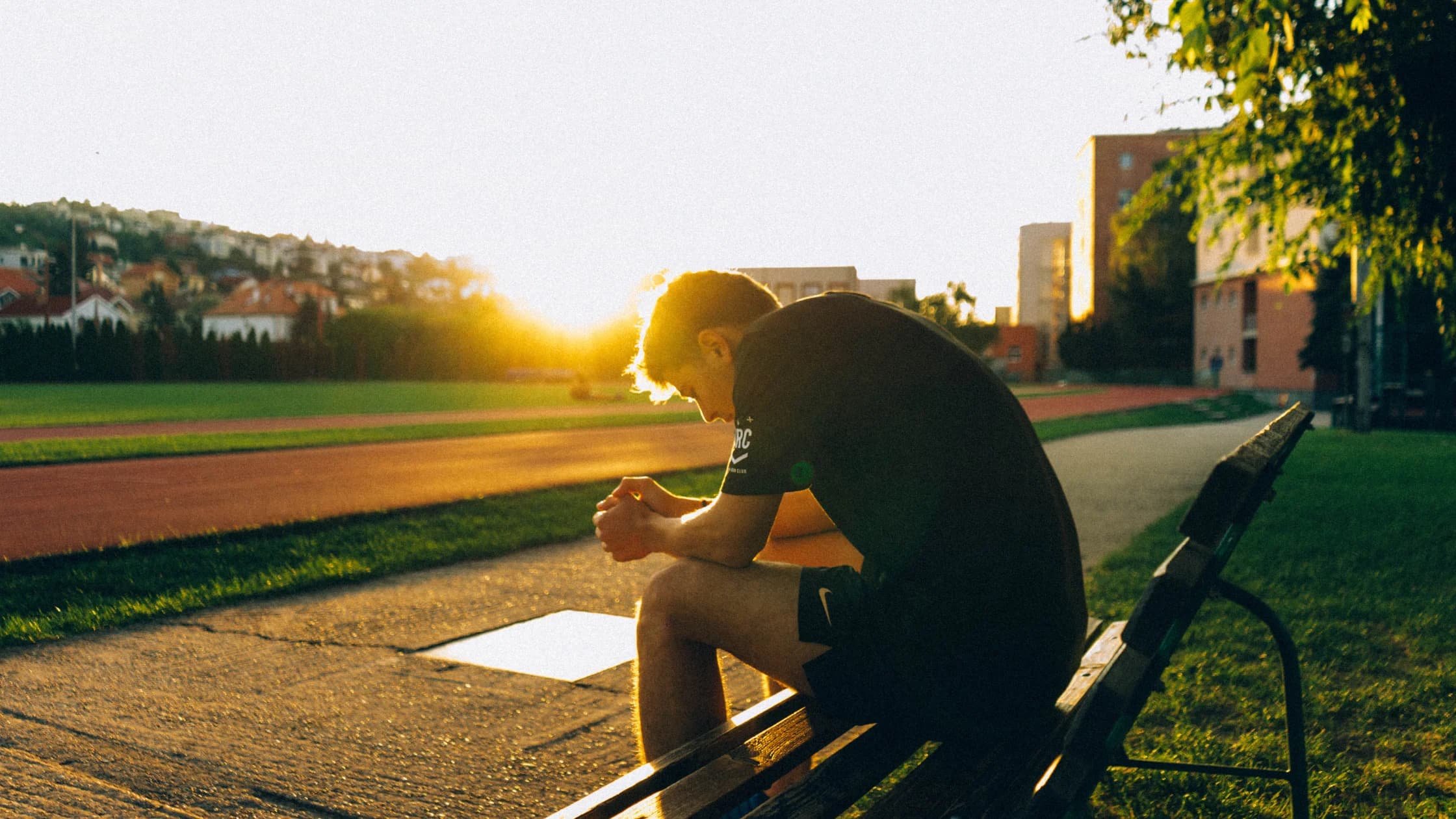 Ein Mann sitzt bei Sonnenuntergang mit gesenktem Kopf und gefalteten Händen auf einer Bank neben einer Laufbahn, als Sinnbild für das Nachdenken über die nächsten Schritte nach dem Ende des Jobs.