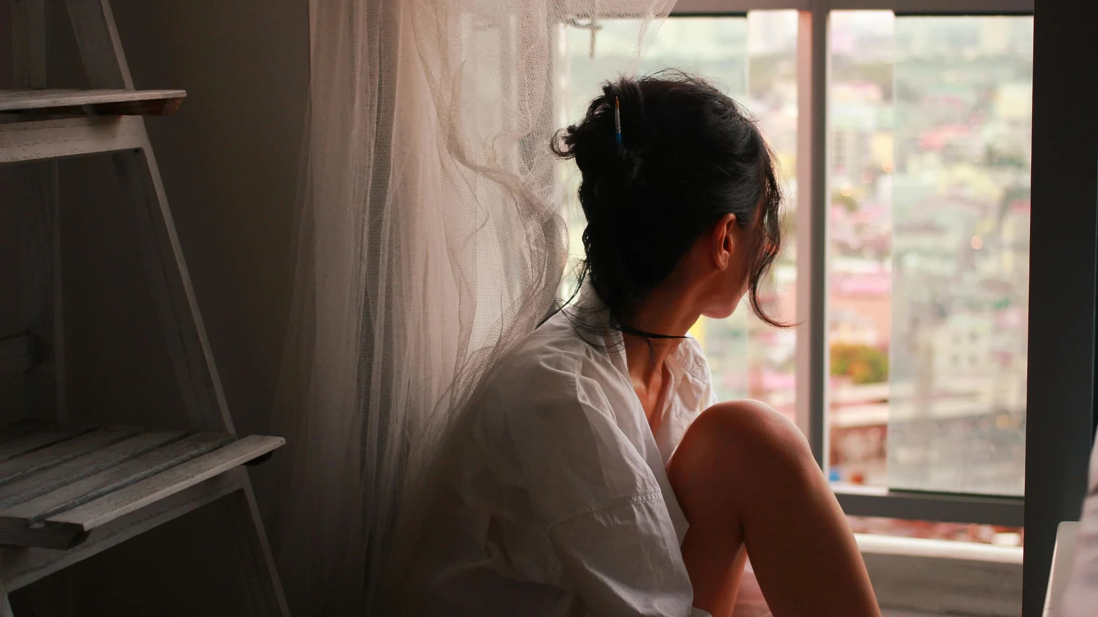 Frau in einem weißen Hemd sitzt am Fenster und blickt nachdenklich auf die Stadt hinaus, während sie über einen beruflichen Neuanfang nach dem Jobverlust nachdenkt