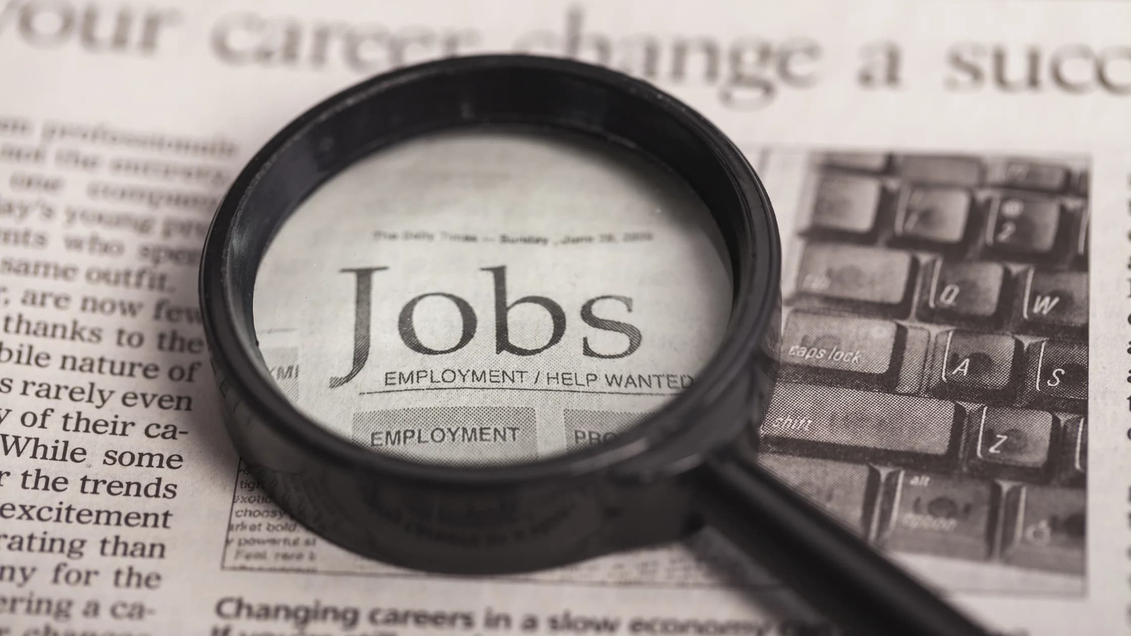 Lupe liegt auf einer Zeitung und vergrößert das Wort „Jobs“ über der Rubrik für Stellenanzeigen, als Sinnbild für die Jobsuche nach der Arbeitslosmeldung