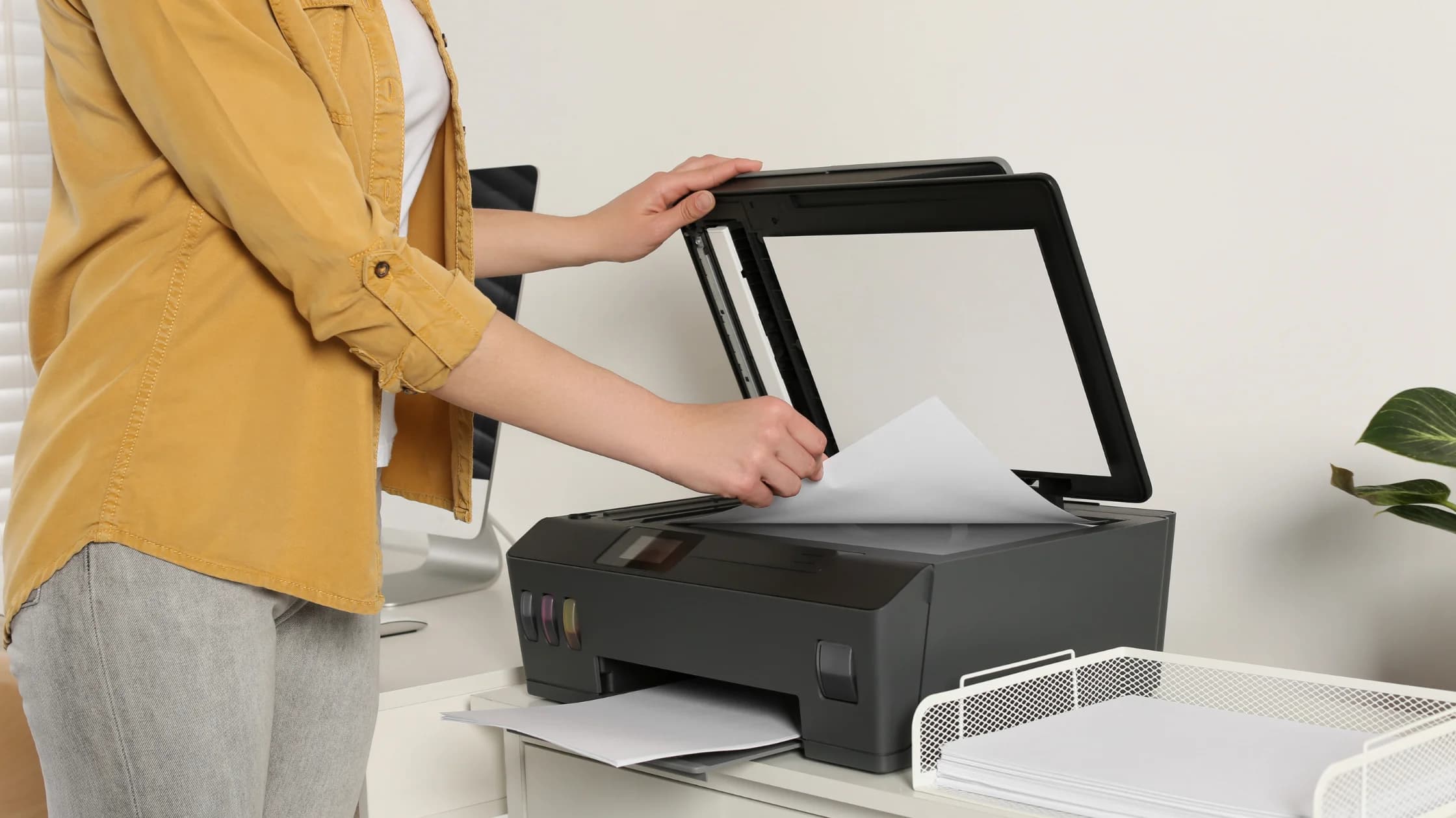 Frau in gelbem Hemd legt ein Blatt Papier auf den Scanner eines Druckers, daneben liegen Papierstapel, als Sinnbild für die Suche nach dem Antrag auf Arbeitslosengeld als PDF