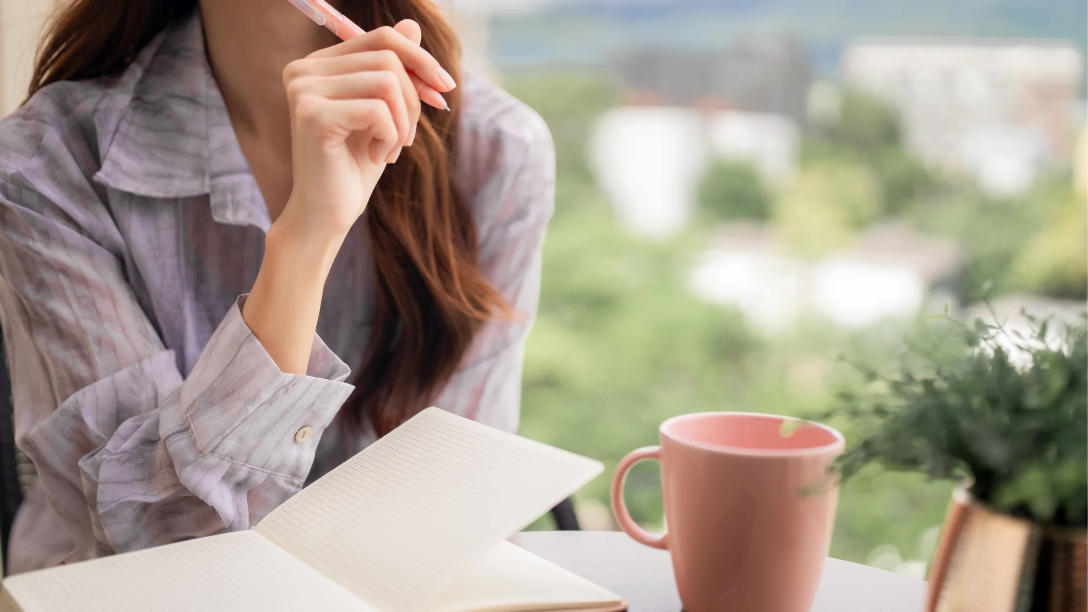 Frau stützt nachdenklich einen Stift ans Kinn, vor ihr liegen ein aufgeschlagenes Notizbuch und eine rosa Tasse auf einem Tisch vor einem Fenster, als Sinnbild für die Planung nach dem Jobverlust