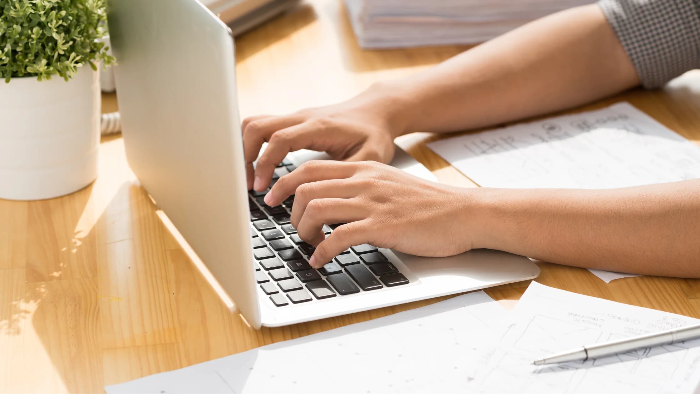 Hände tippen auf der Tastatur eines silbernen Laptops auf einem Holztisch, daneben liegen Papiere mit Skizzen und ein Stift, als Sinnbild für den Online-Antrag auf Arbeitslosengeld