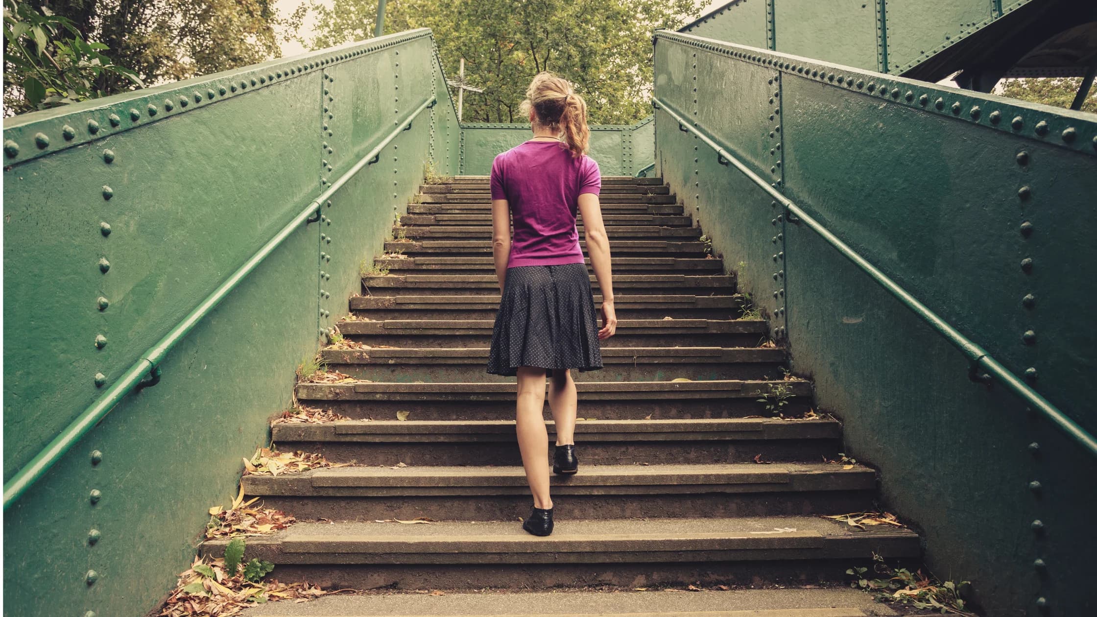Eine Frau geht von hinten gesehen eine breite Treppe mit grünen Geländern im Freien hinauf, als Sinnbild für einen Neustart nach dem Ende des Arbeitsverhältnisses.
