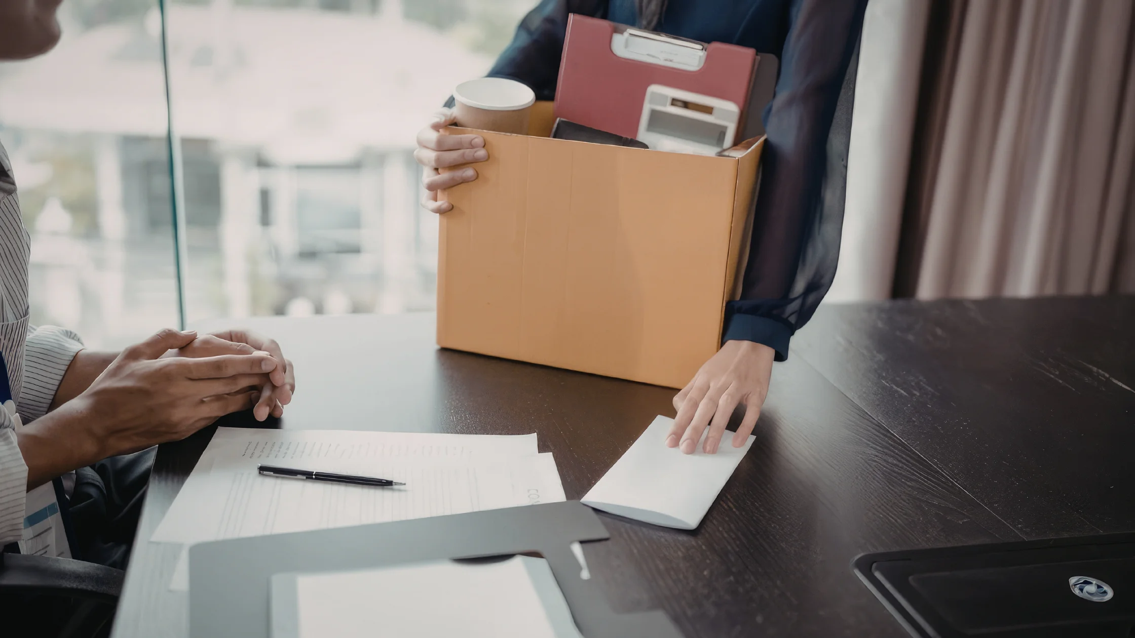 Eine Person sitzt im Büro und hält einen Karton mit ihren persönlichen Sachen, gegenüber sitzt eine zweite Person mit gefalteten Händen, auf dem Tisch liegen Papiere und ein Umschlag, als Sinnbild für ein Kündigungsgespräch.