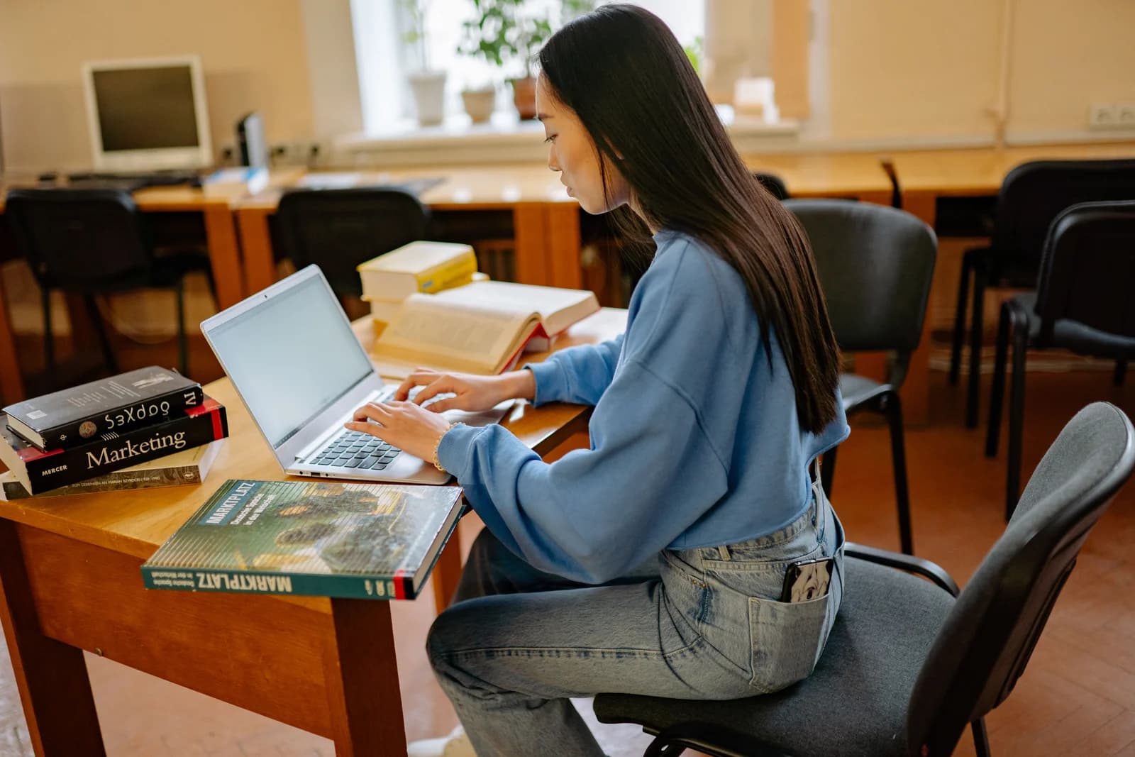 Eine Frau sitzt in einem Raum mit Holztischen an einem Laptop, neben ihr liegen aufgeschlagene und gestapelte Bücher.