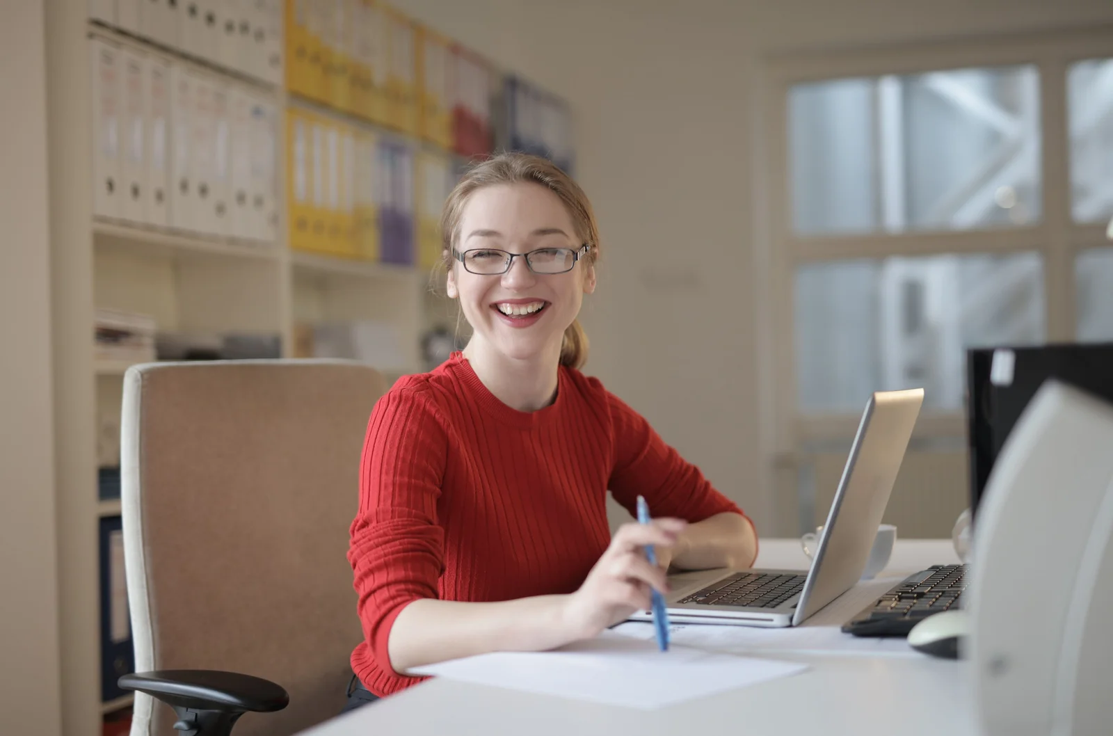 Eine lachende Frau mit Brille und rotem Pullover sitzt mit Stift und Papier an einem Schreibtisch vor einem Laptop.