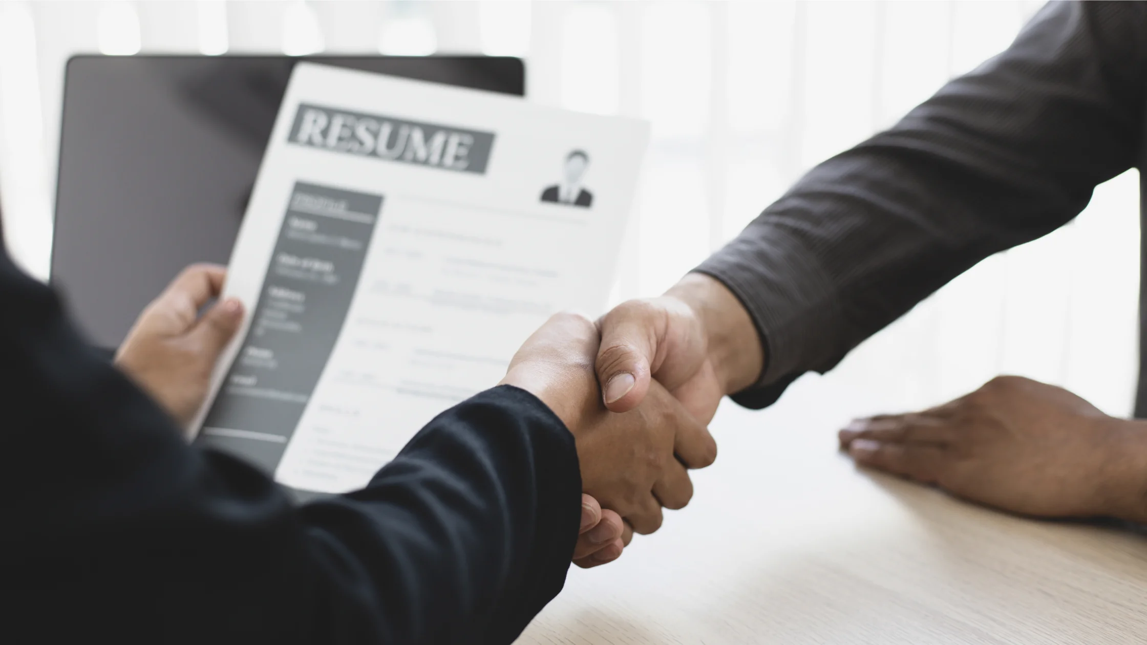 Zwei Personen geben sich über einem Tisch die Hand, eine von ihnen hält einen Lebenslauf in der anderen Hand, als Sinnbild für das Gespräch bei der Agentur für Arbeit